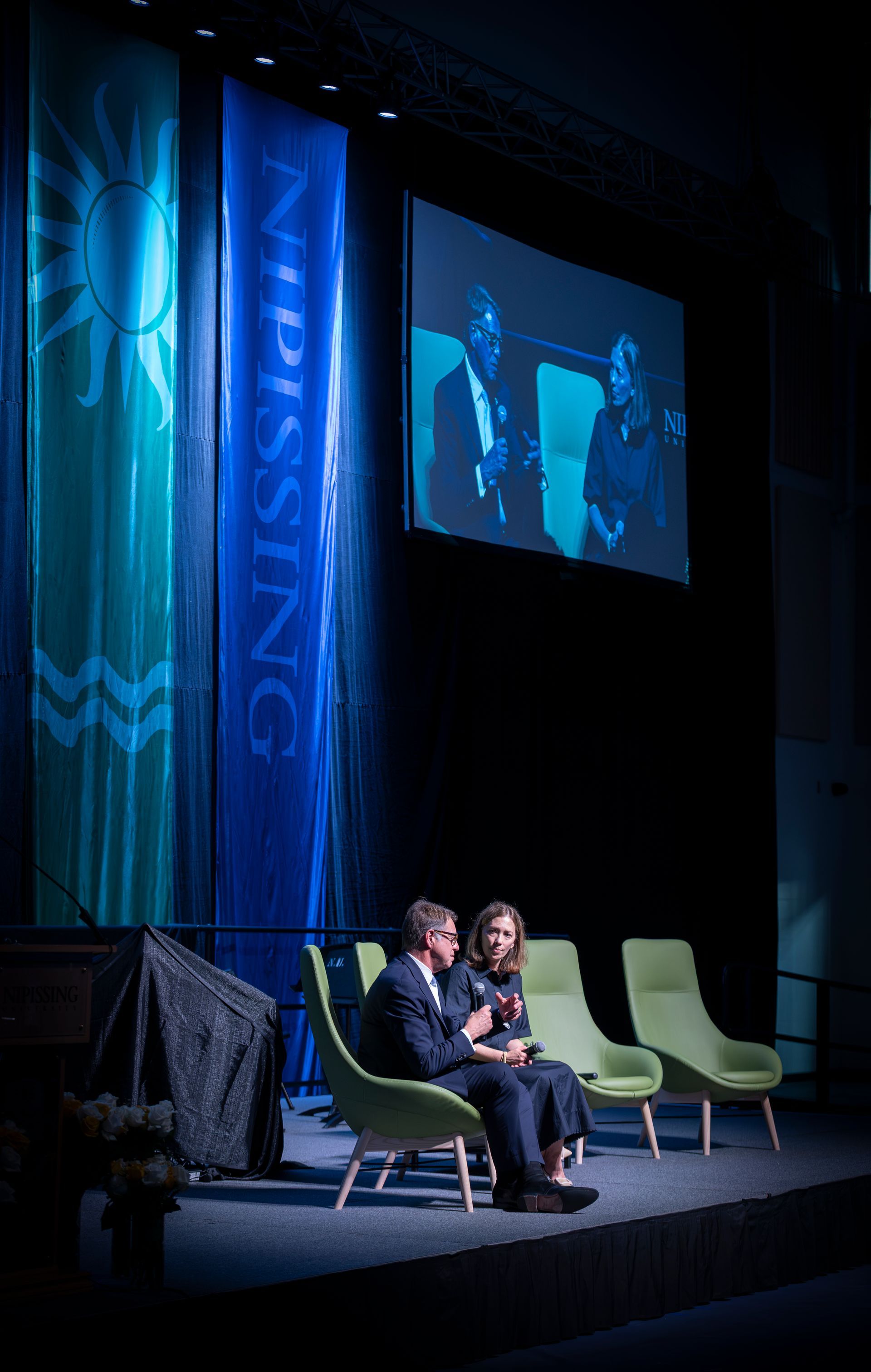 Two people seated on stage during a panel, with blue-lit backdrop and empty chairs nearby.