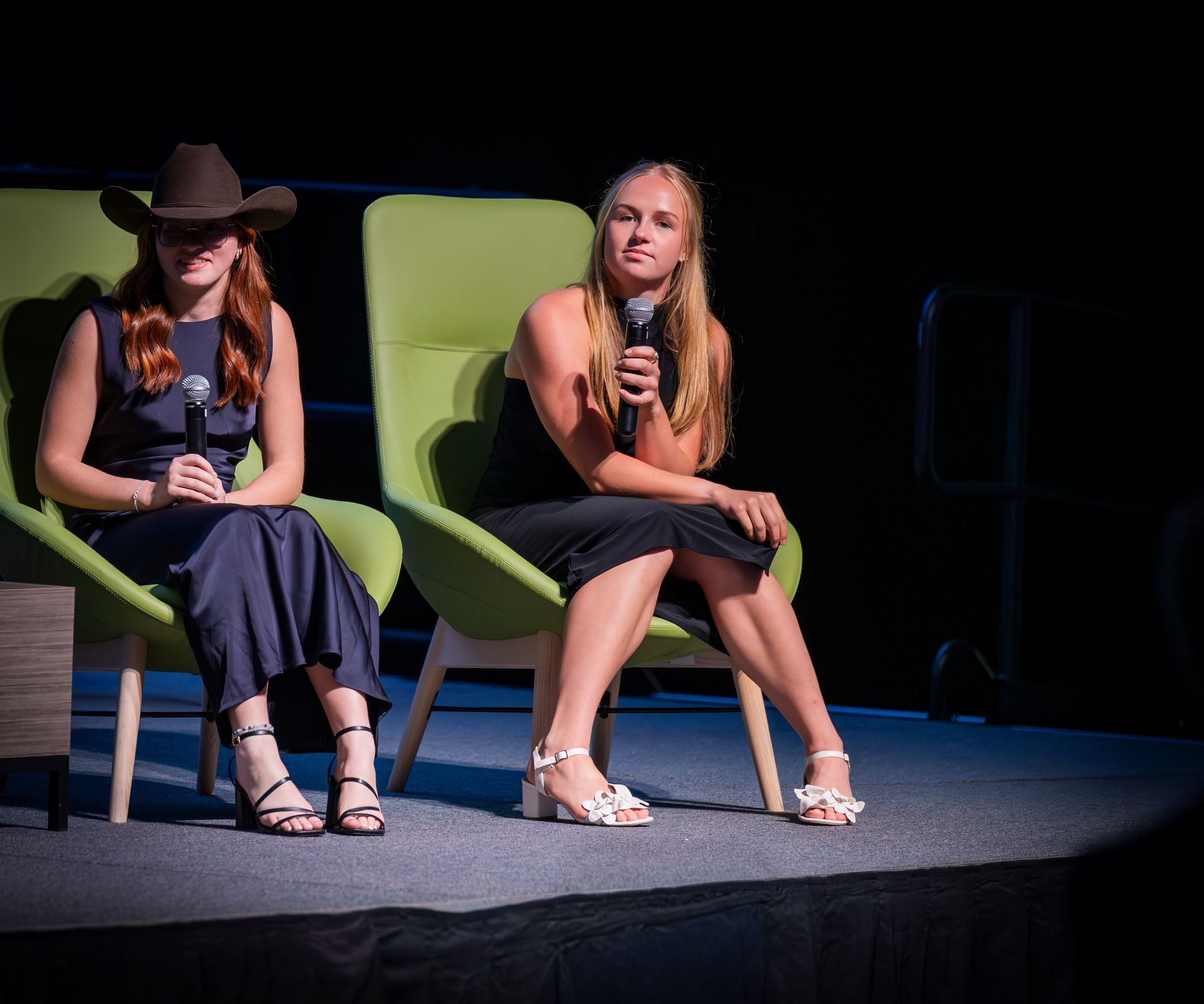 Two women seated on stage holding microphones, one in a cowboy hat, under spotlighting