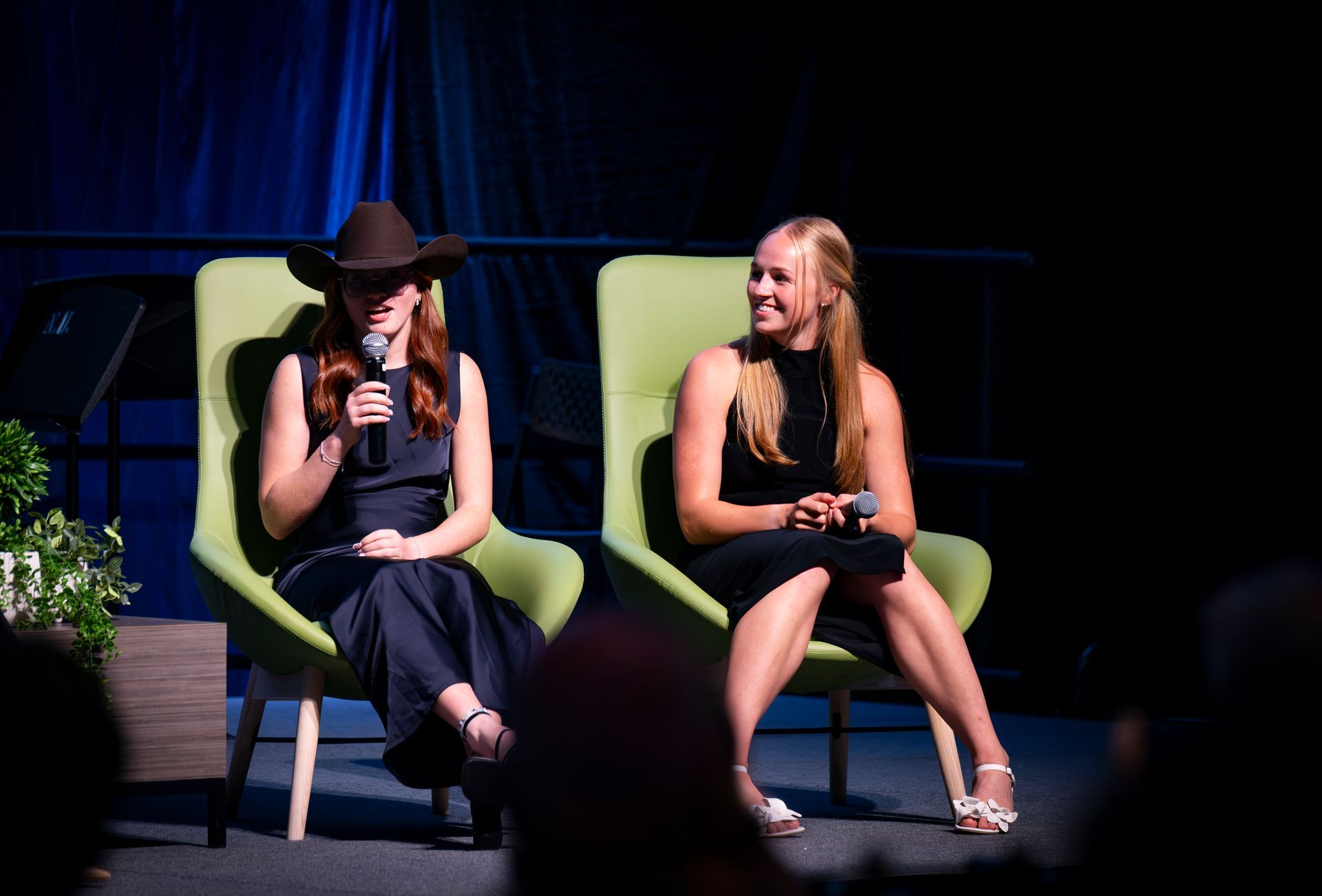 Two women on stage in green chairs, one speaking into a microphone, one smiling beside her.