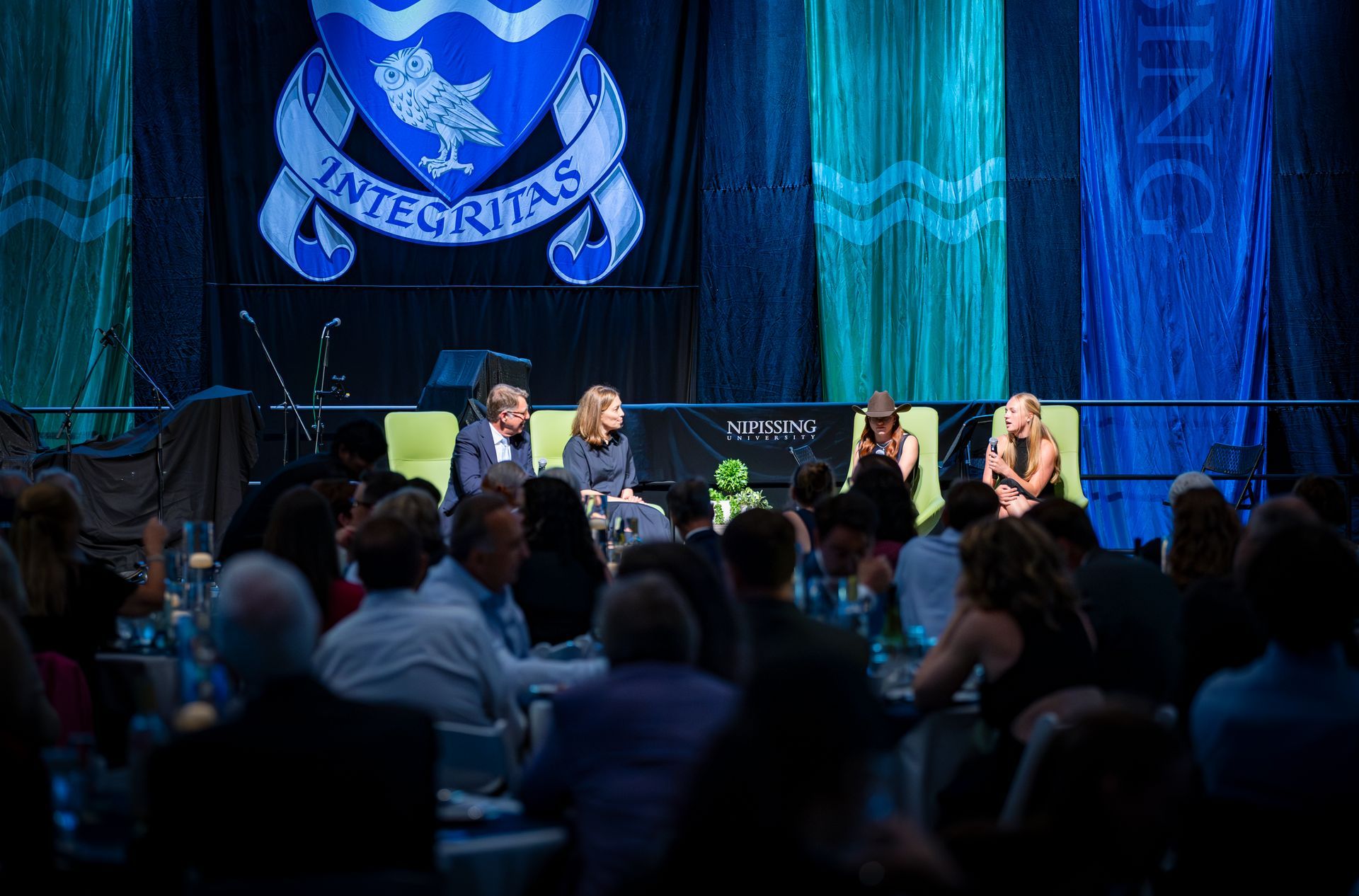 Panel discussion on a blue-lit stage with seated speakers and a large crest backdrop