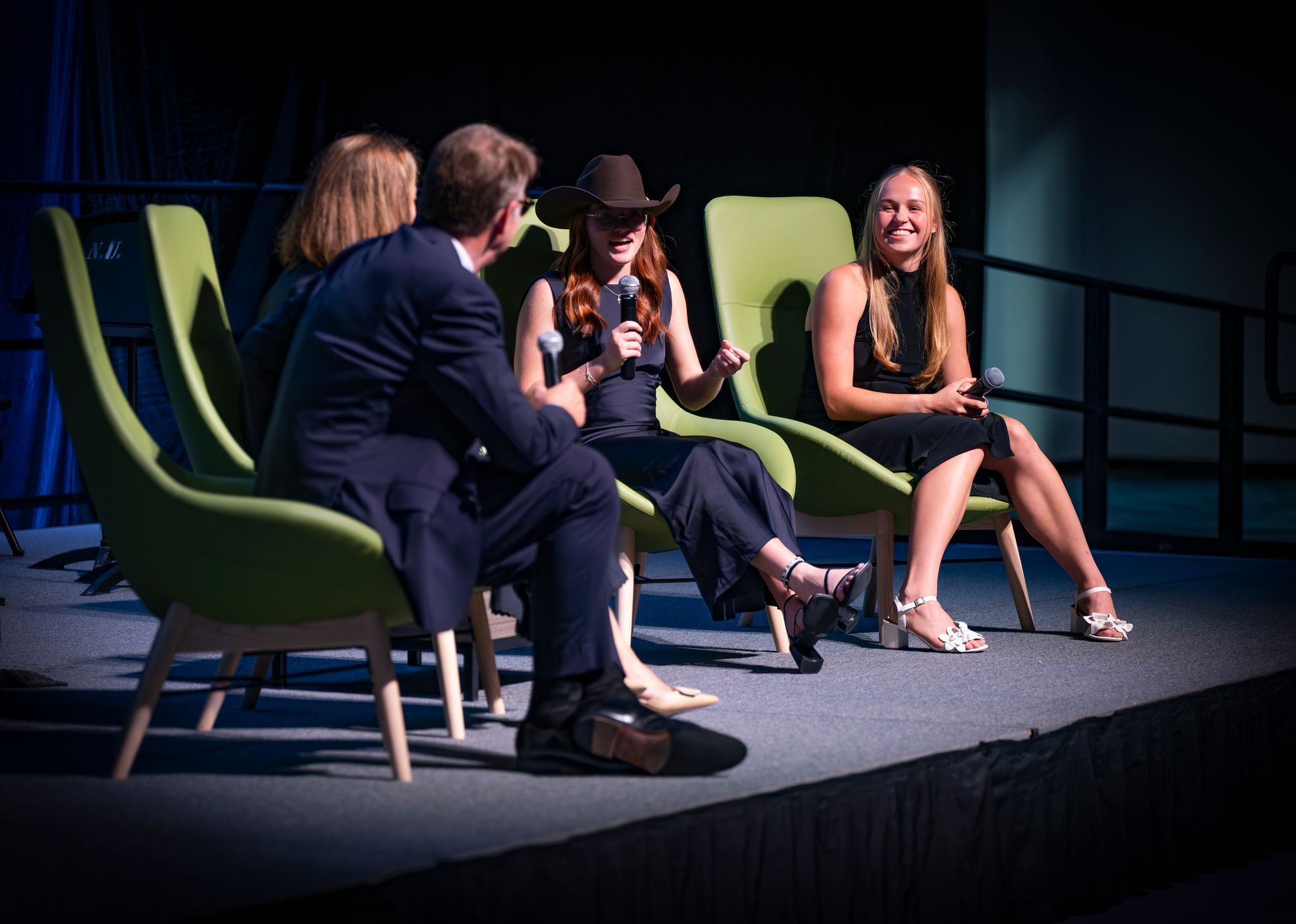 Four people seated on stage in green chairs, one speaking into a microphone, under spotlight lighting.