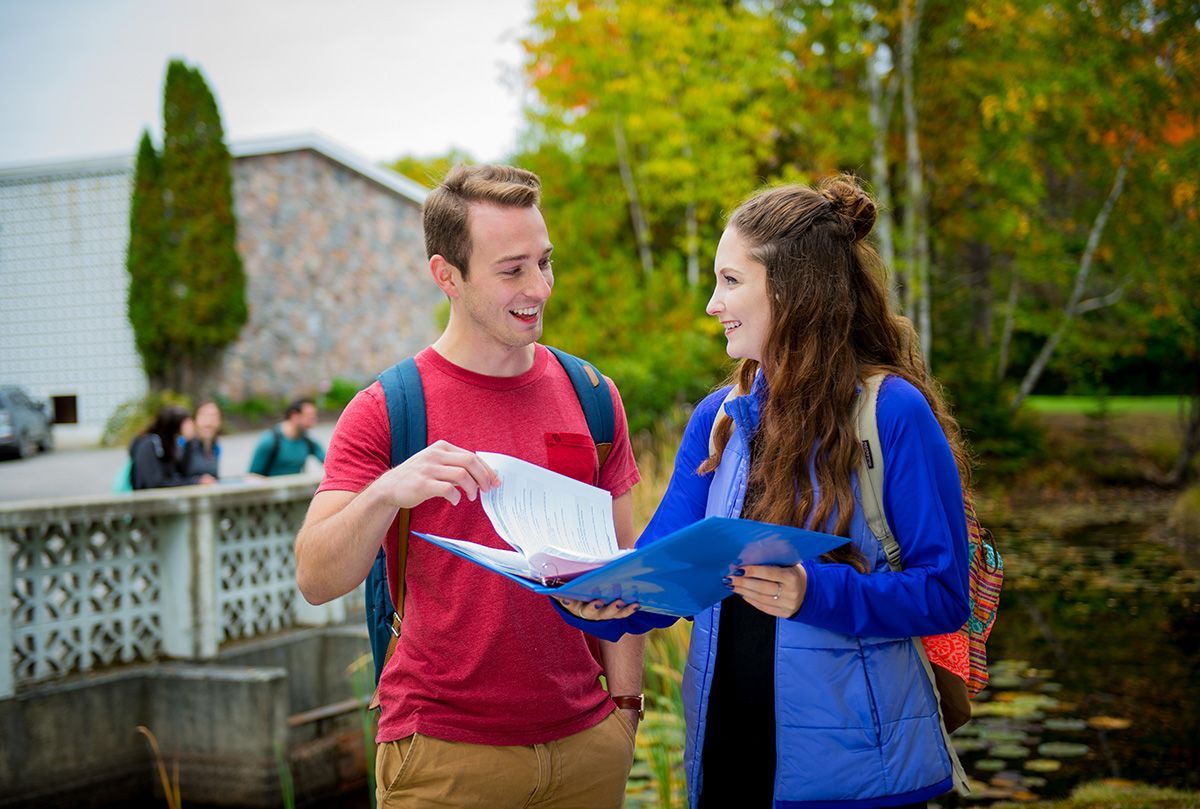 Two students outdoors, smiling and holding papers. One in red shirt, one in blue jacket, near a pond.
