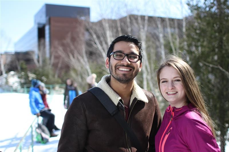 Two smiling people outside on a snowy day. Nipissing University building in the backgrou8nd.