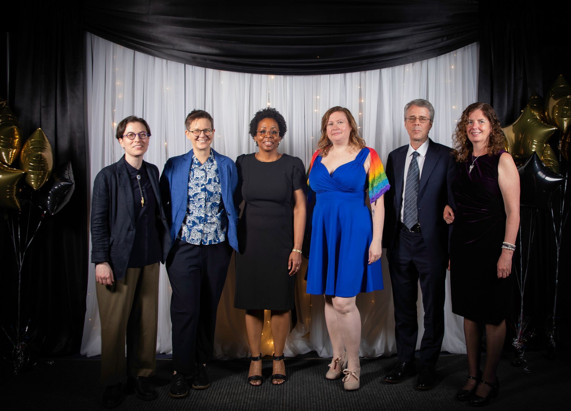 Six people posing for a formal group photo in front of a black and white backdrop, some with formal attire.