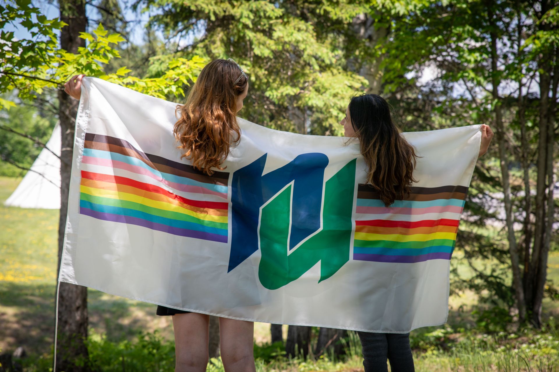 Two people hold a white flag with a rainbow stripe, Nipissing University logo, and nature setting.