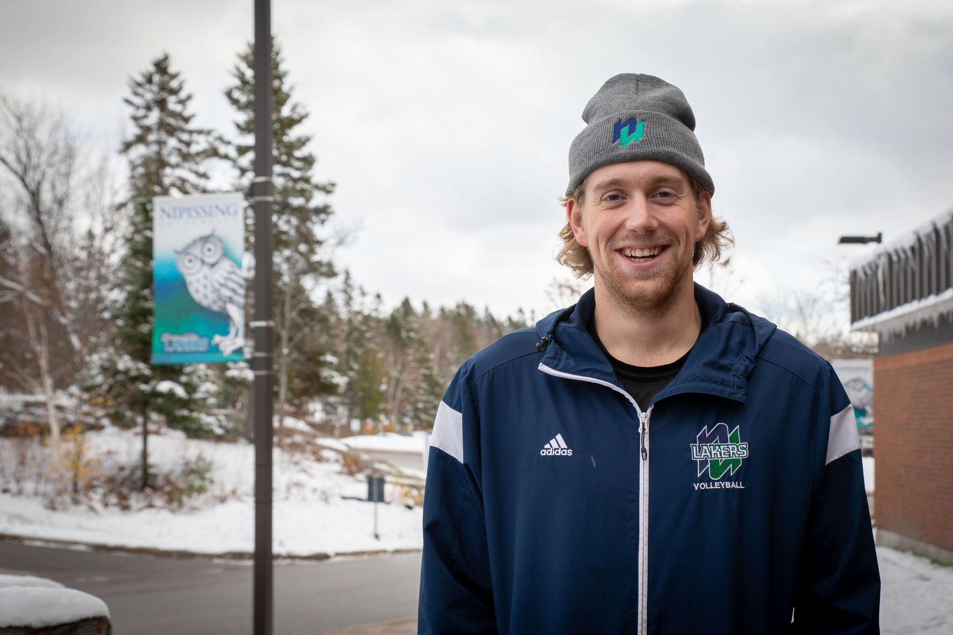 Man smiling, wearing a beanie and jacket outdoors in a snowy setting with a banner in the background.