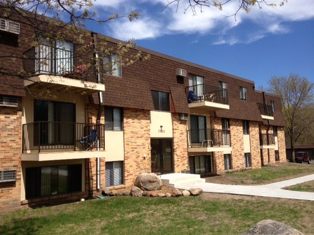 Apartment building with brown roof, brick exterior, and balconies. Blue sky with clouds and grassy lawn.