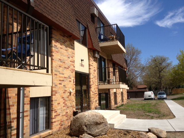 Apartment building with brick exterior, balconies, and sidewalk. Sunny day with blue sky.