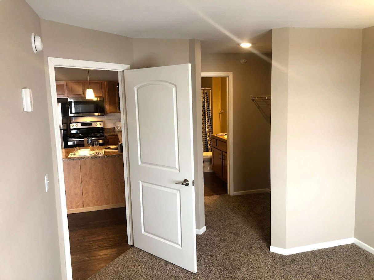 Doorway to a kitchen and bathroom. Beige walls, brown carpet, white door.