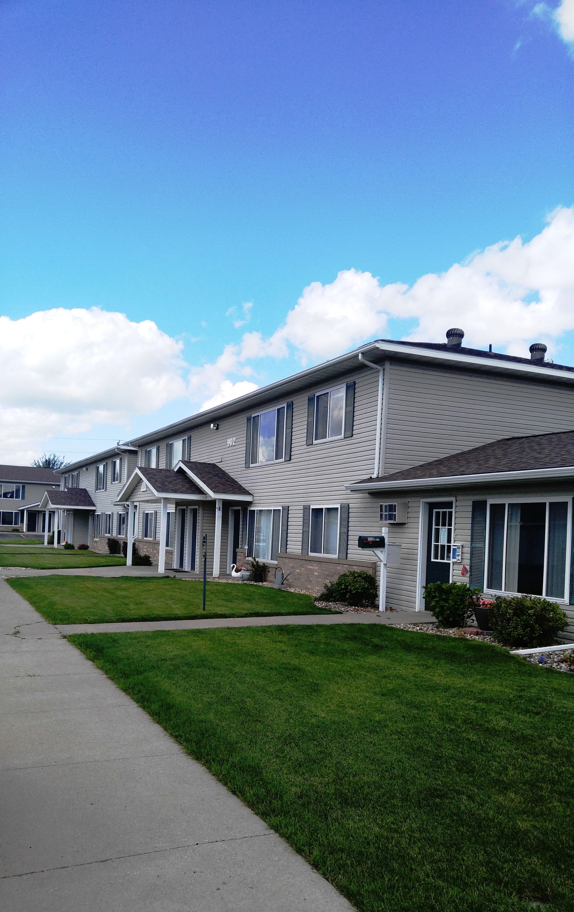 Row of light-colored townhouses with green lawns and a sidewalk under a blue sky with clouds.