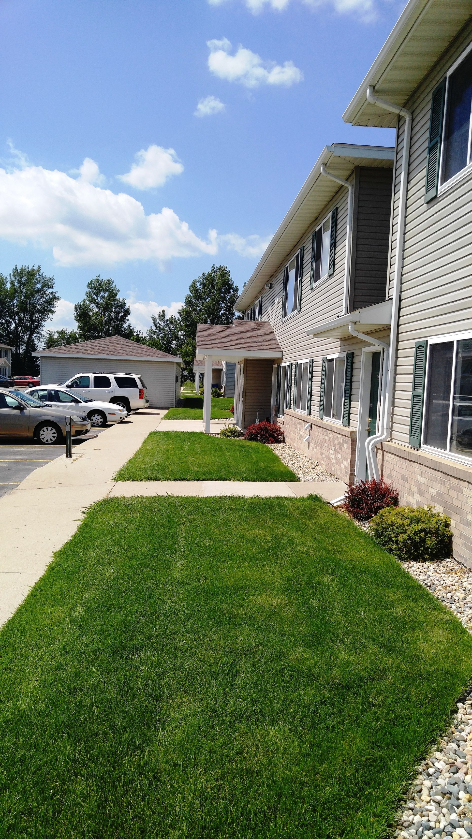 Apartment building with green lawns, blue sky. Cars parked along a walkway.