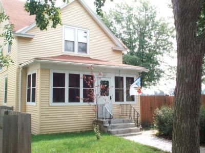 Yellow two-story house with a porch, steps, and a flag; green lawn and trees in front.