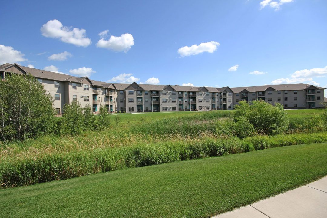 Apartment complex on a grassy hill, with a wetland in the foreground, under a blue sky with clouds.