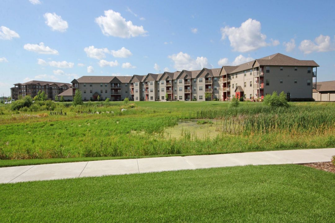 Apartment buildings line a green field under a blue sky with clouds. A sidewalk and grass in the foreground.