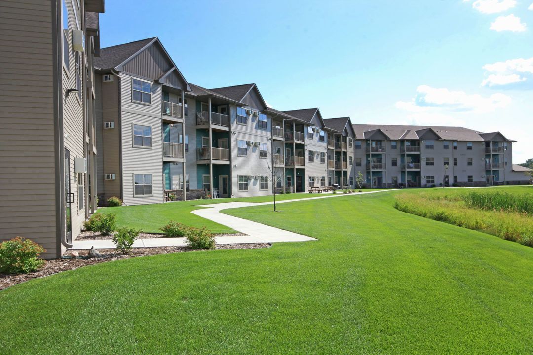 Apartment buildings with balconies, on a sunny day. Green grass and a paved path in front.