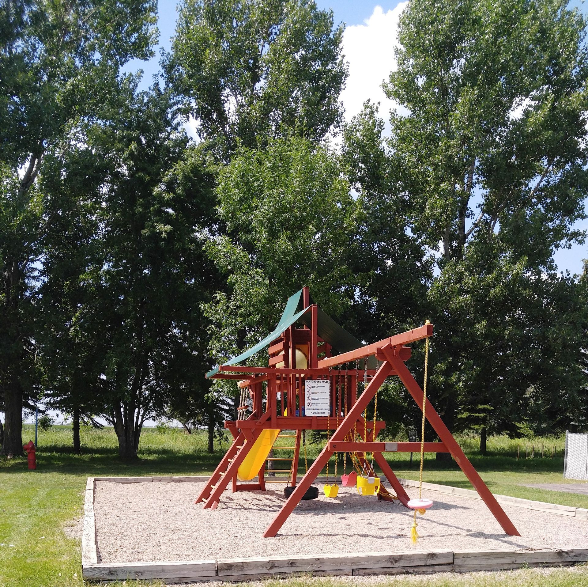 Playground with red wooden structure, swings, and a slide, set against a backdrop of green trees and a blue sky.