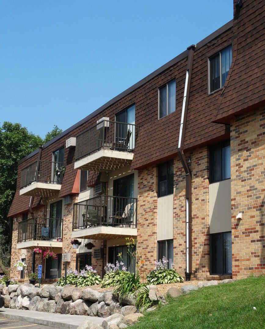 Apartment building with brick facade, brown roof, balconies, and landscaping under a blue sky.