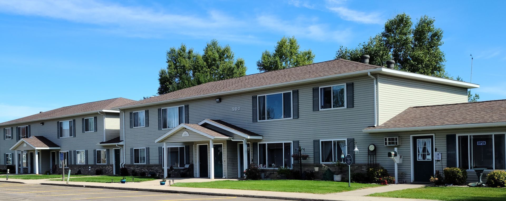Apartment complex with gray siding and brown roofs under a blue sky, with green grass and trees.