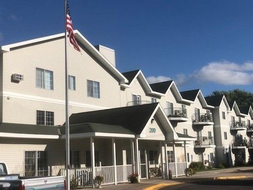 Beige multi-story assisted living facility with flag. Black roof and balconies. Blue sky.