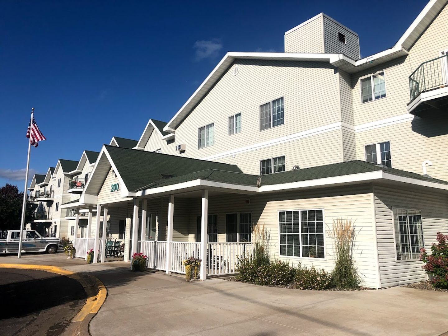 Senior living facility with American flag and blue sky. Beige siding and green roof.