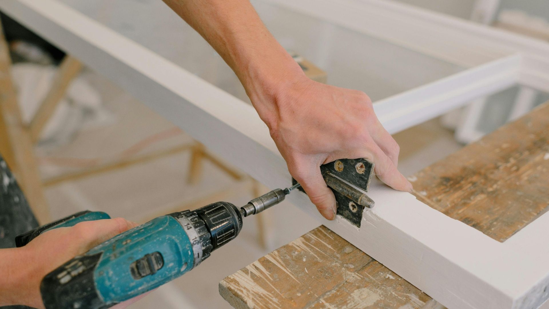 Person using a power drill to attach a hinge to a white painted window frame.