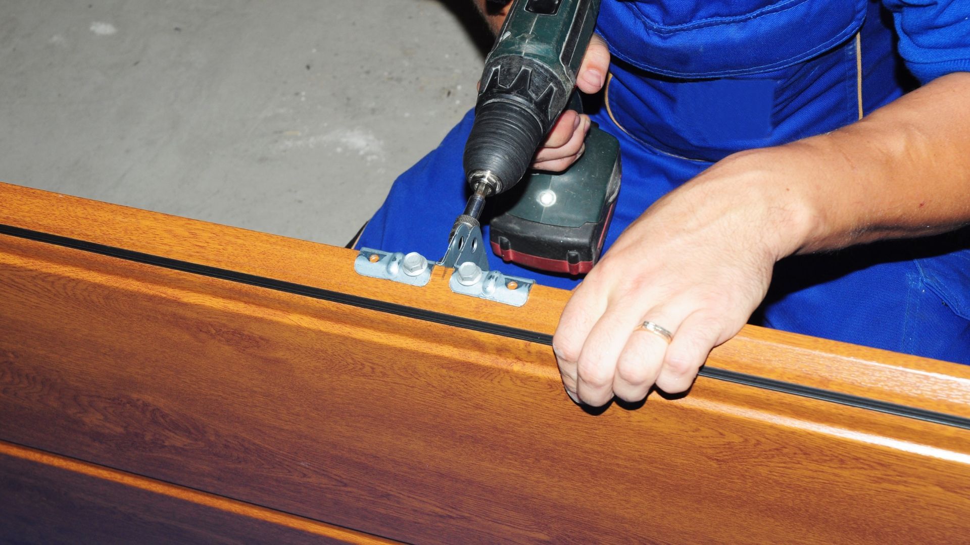 Person in blue overalls using a power drill to attach hinges to a wooden frame.
