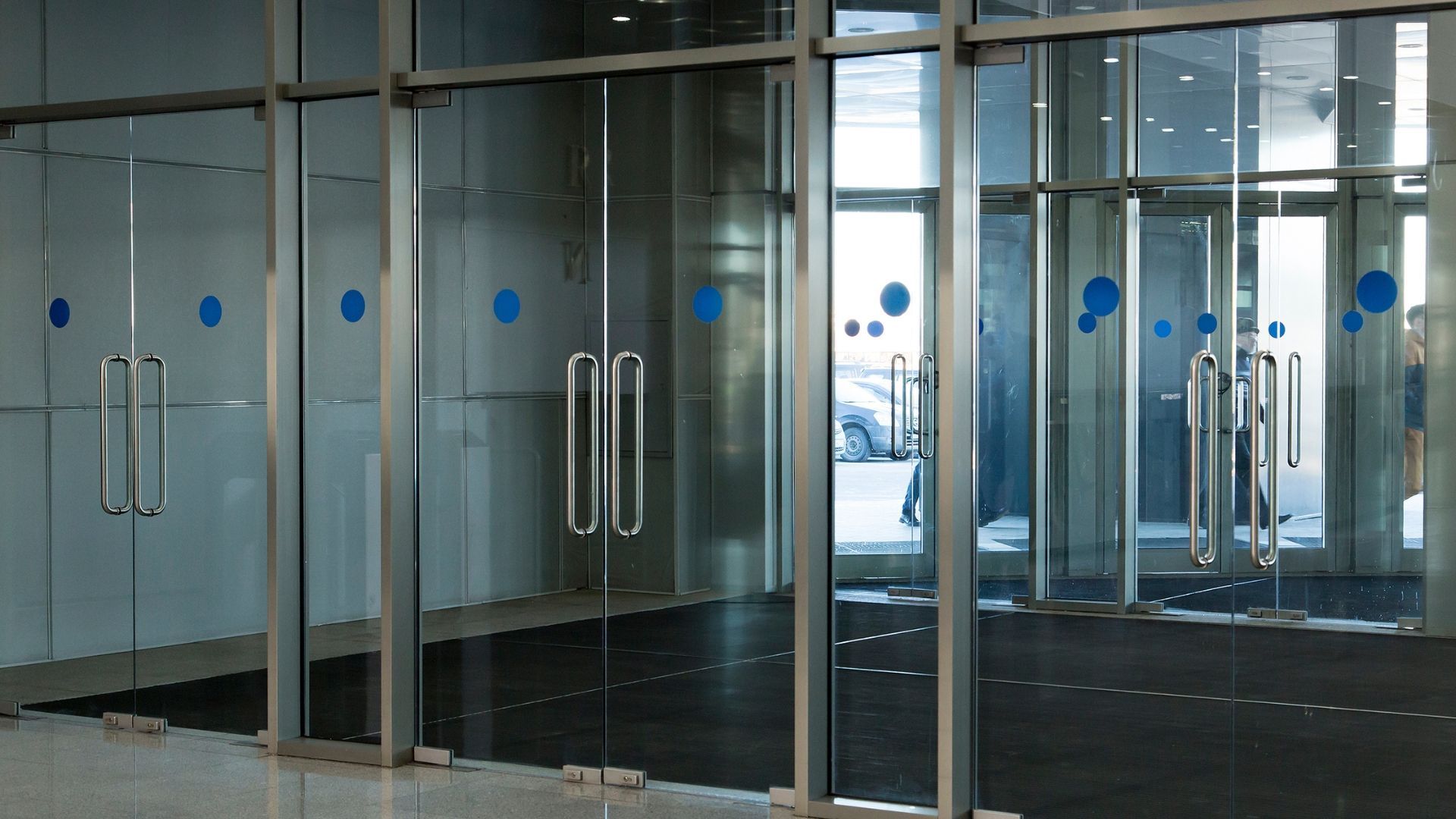 Glass doors of a building with blue dot stickers and chrome handles; a person is visible through the doors.