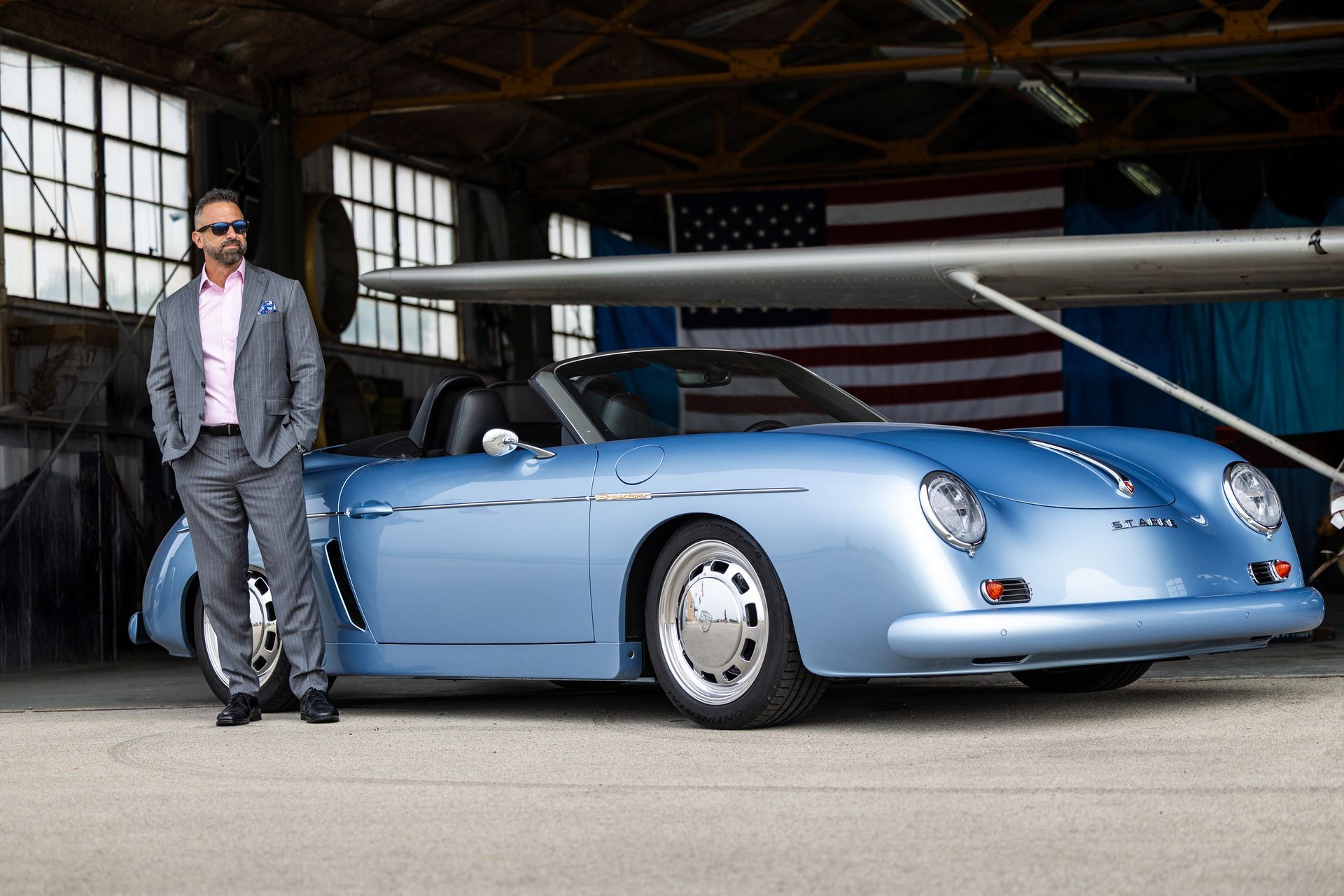 Man in a gray suit standing beside a light blue vintage convertible in an airplane hangar