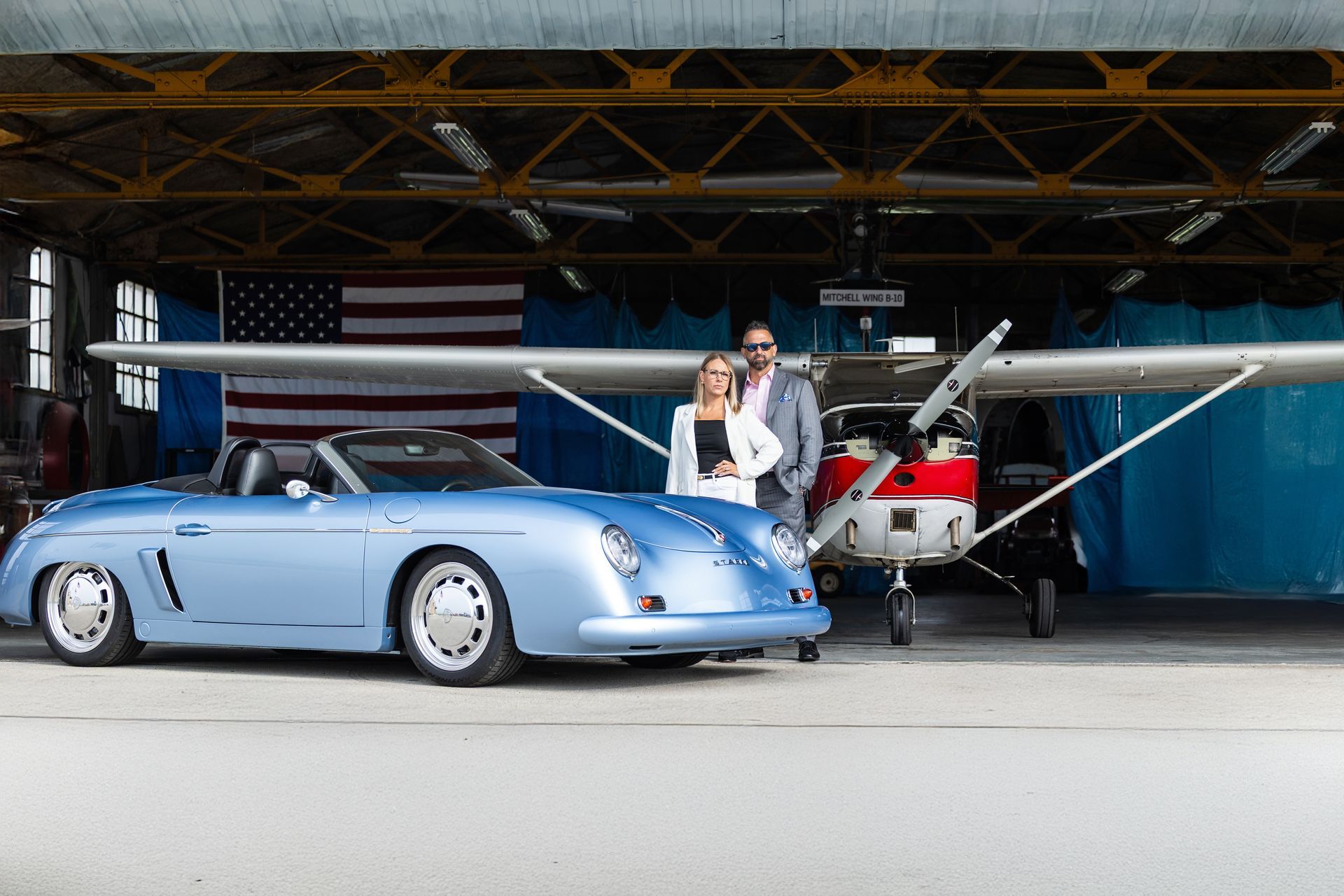 Classic light-blue sports car and small plane in a hangar, with a person standing beside them.