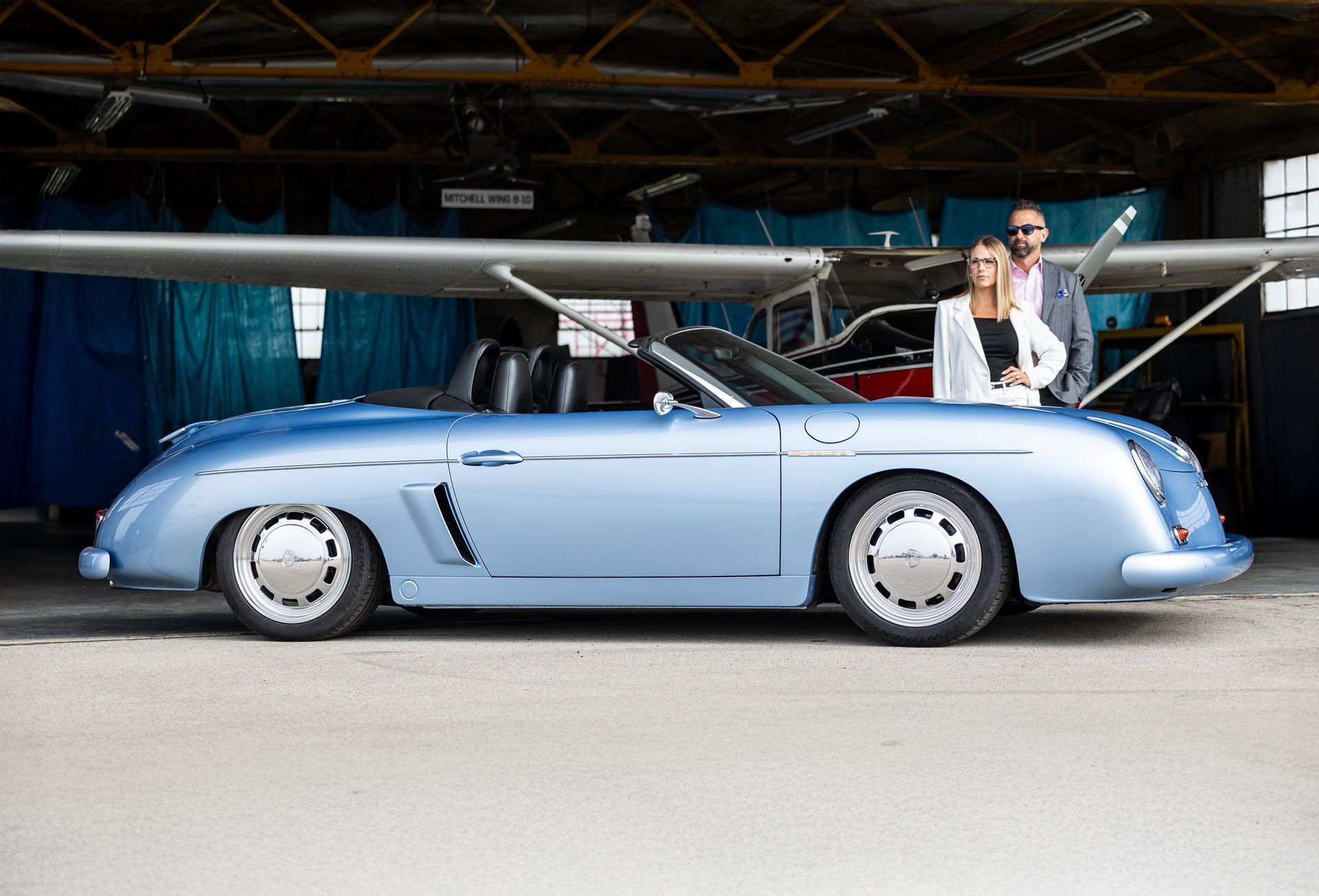 Light blue vintage car in a hangar with a man standing beside it and a small airplane behind.