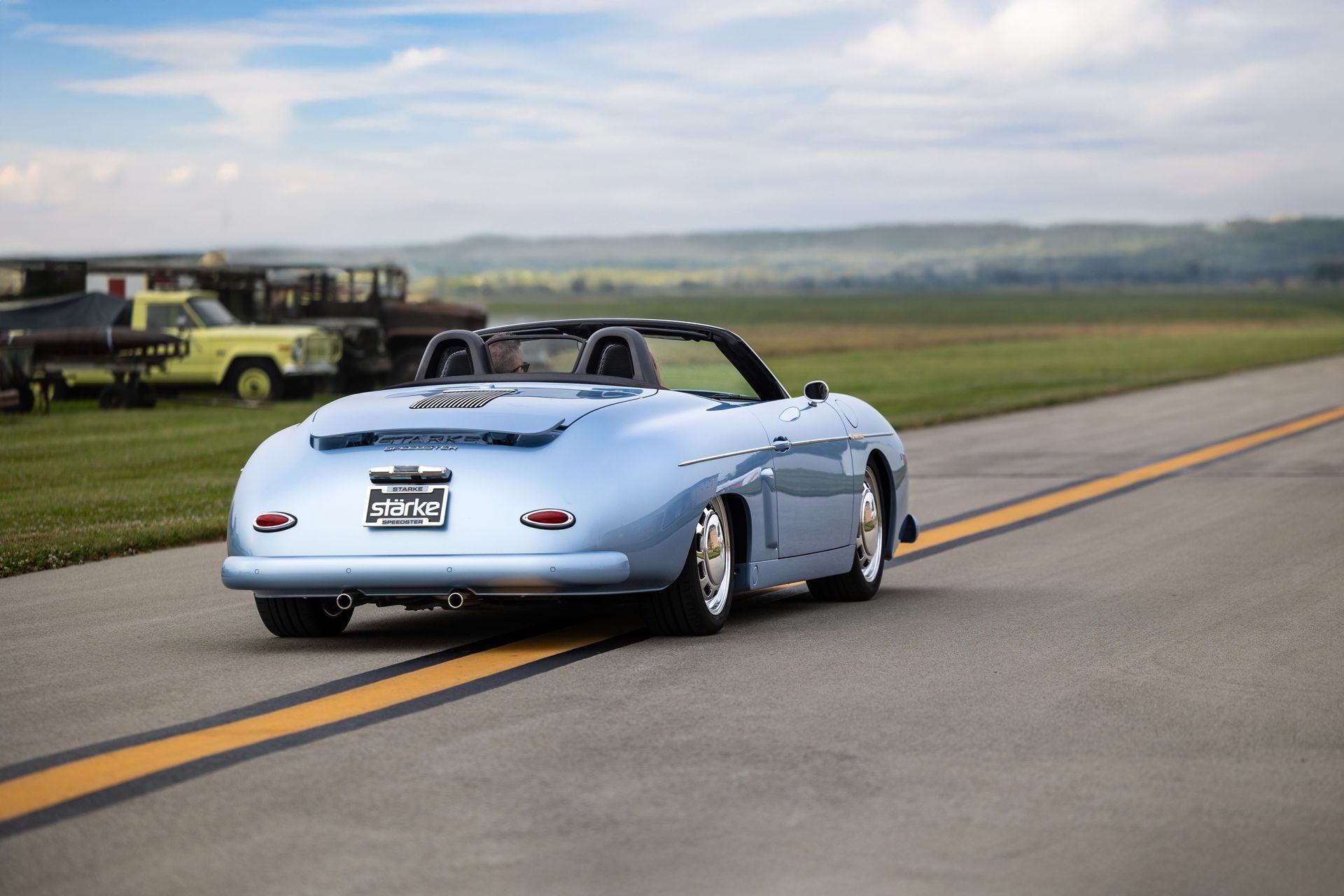 Light blue convertible sports car driving on a rural road with parked vehicles in the background