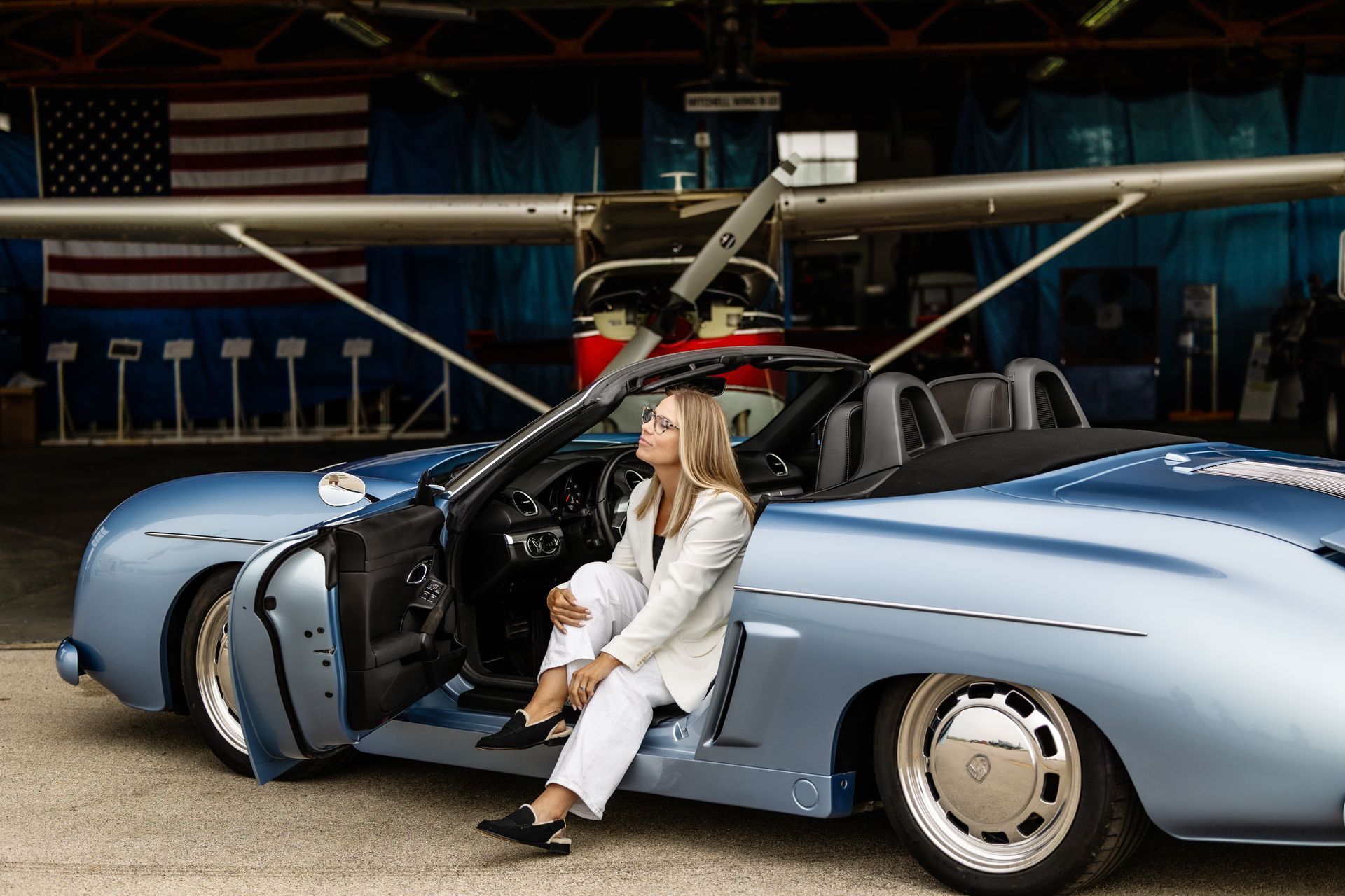 Woman sitting by a light blue sports car with its door open in a hangar, with a helicopter behind it
