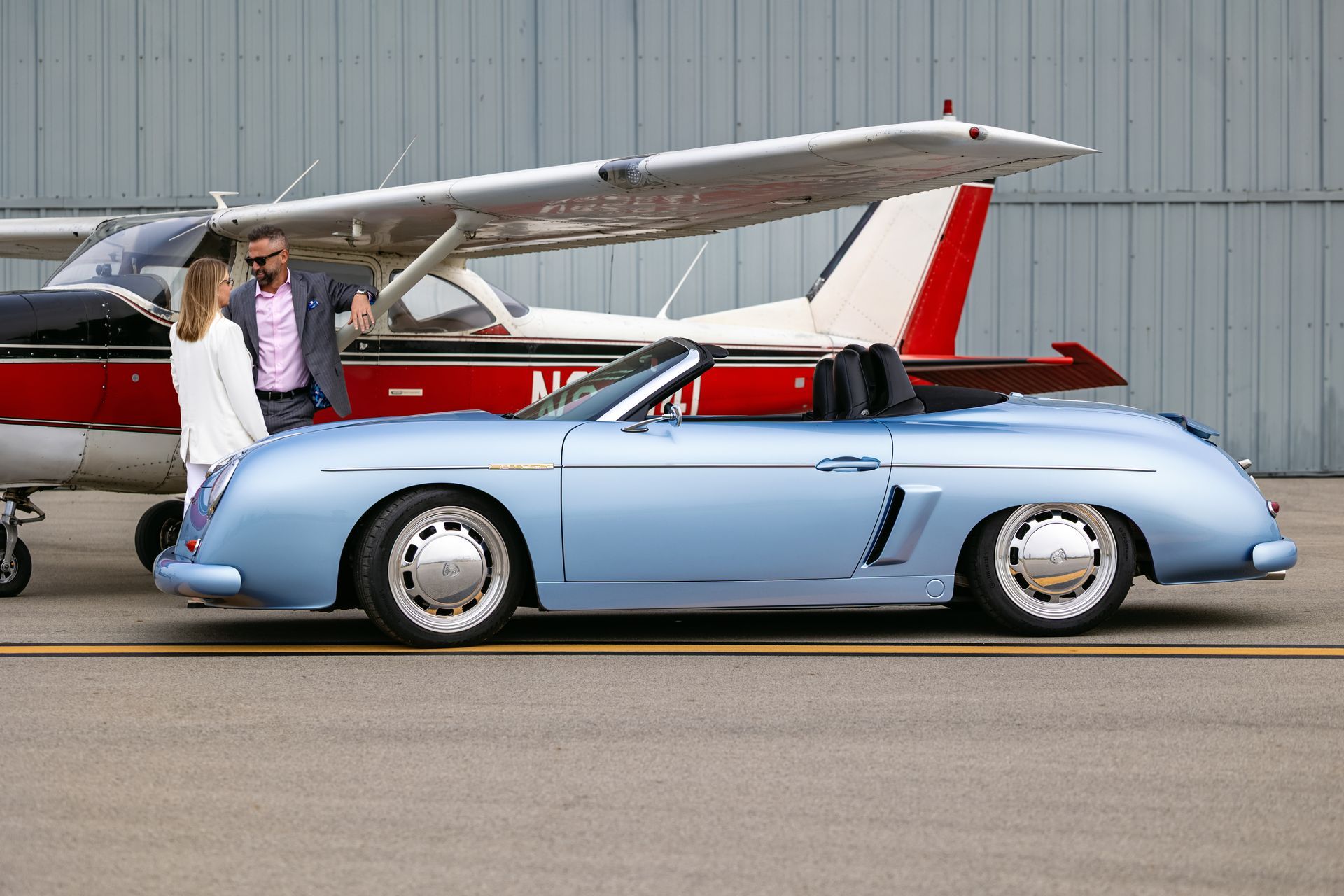 Two people by a light blue vintage convertible beside a small airplane on a runway
