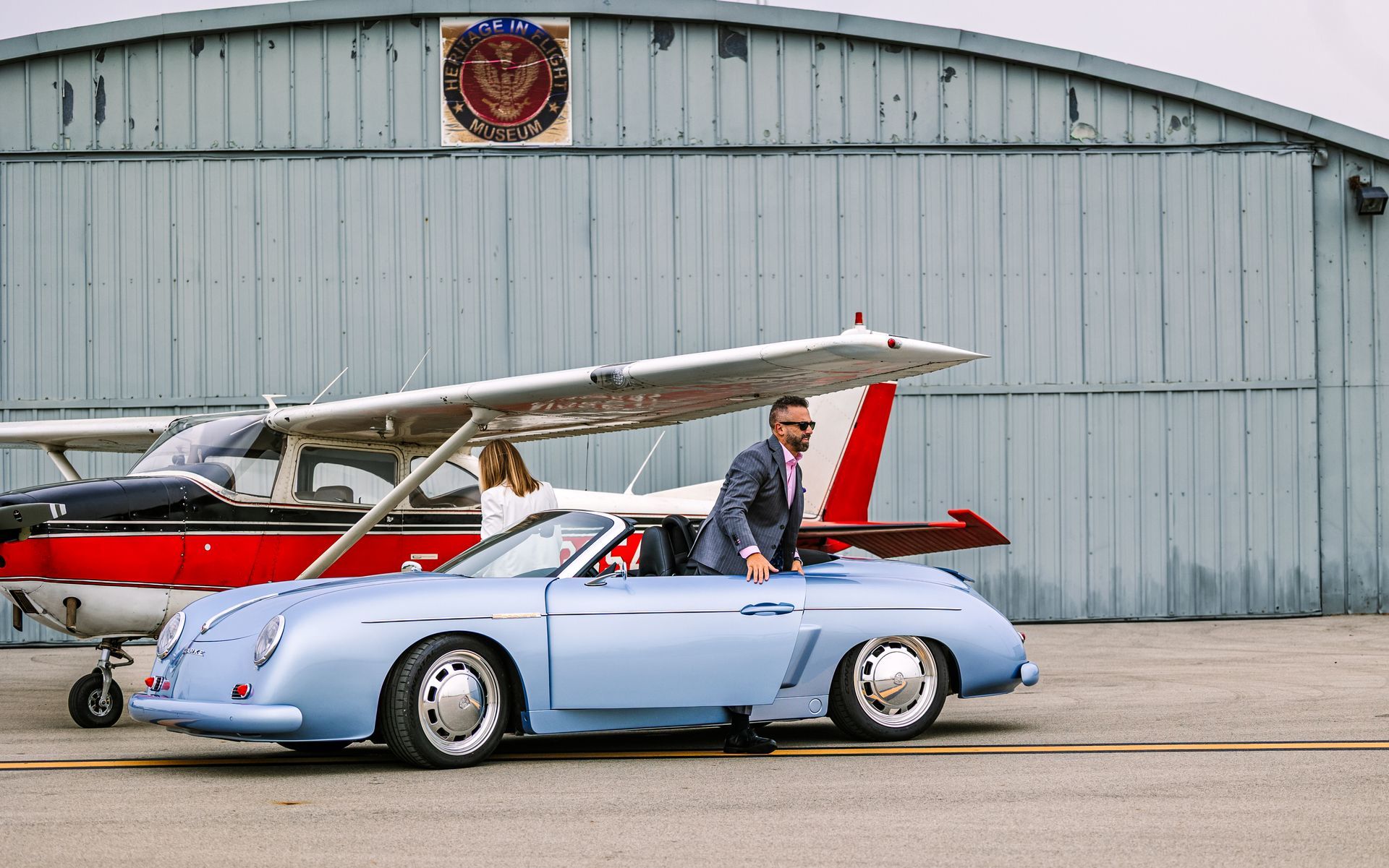 Light blue convertible beside a small red-and-white airplane outside a hangar