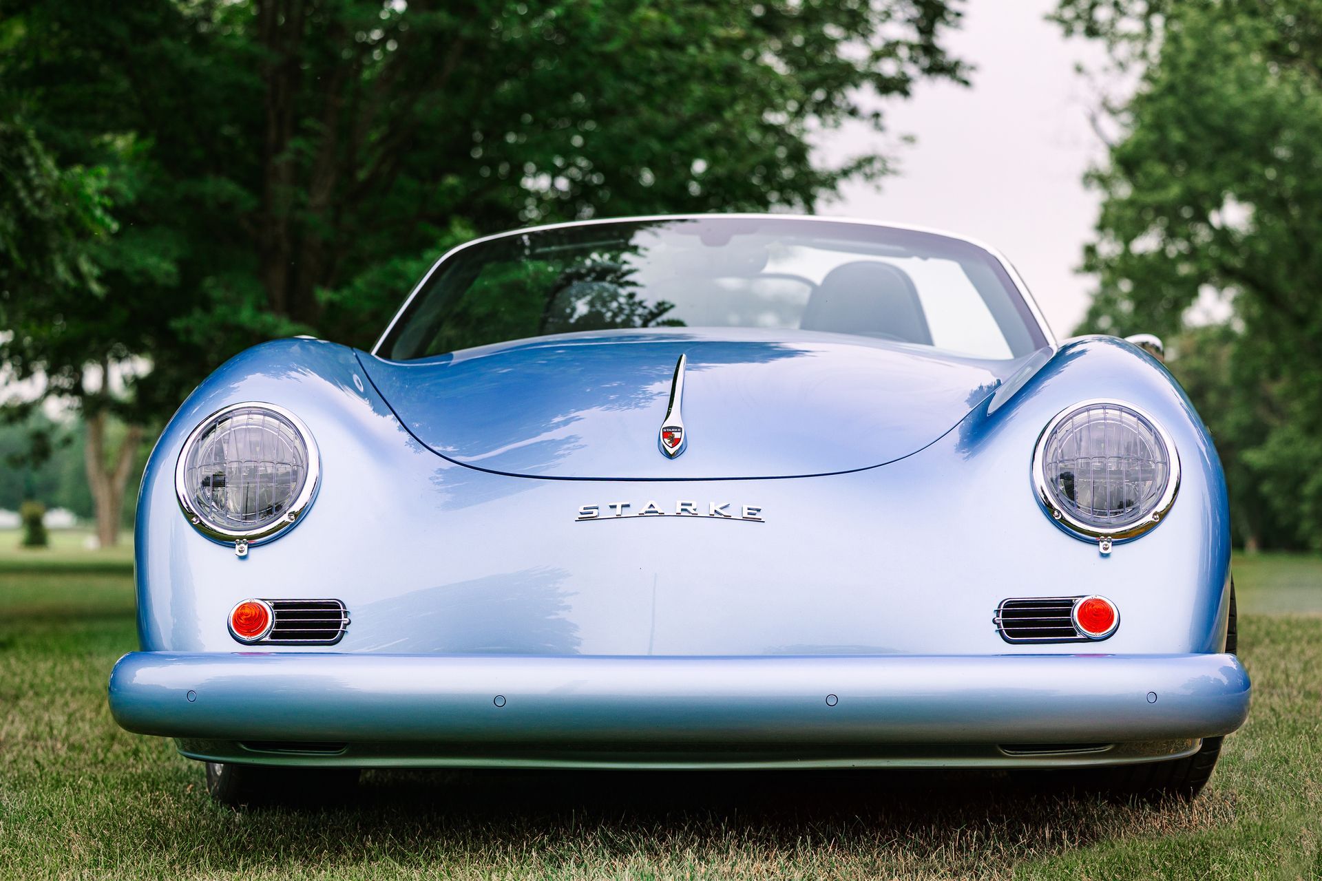 Front view of a light blue classic sports car parked on grass with trees in the background