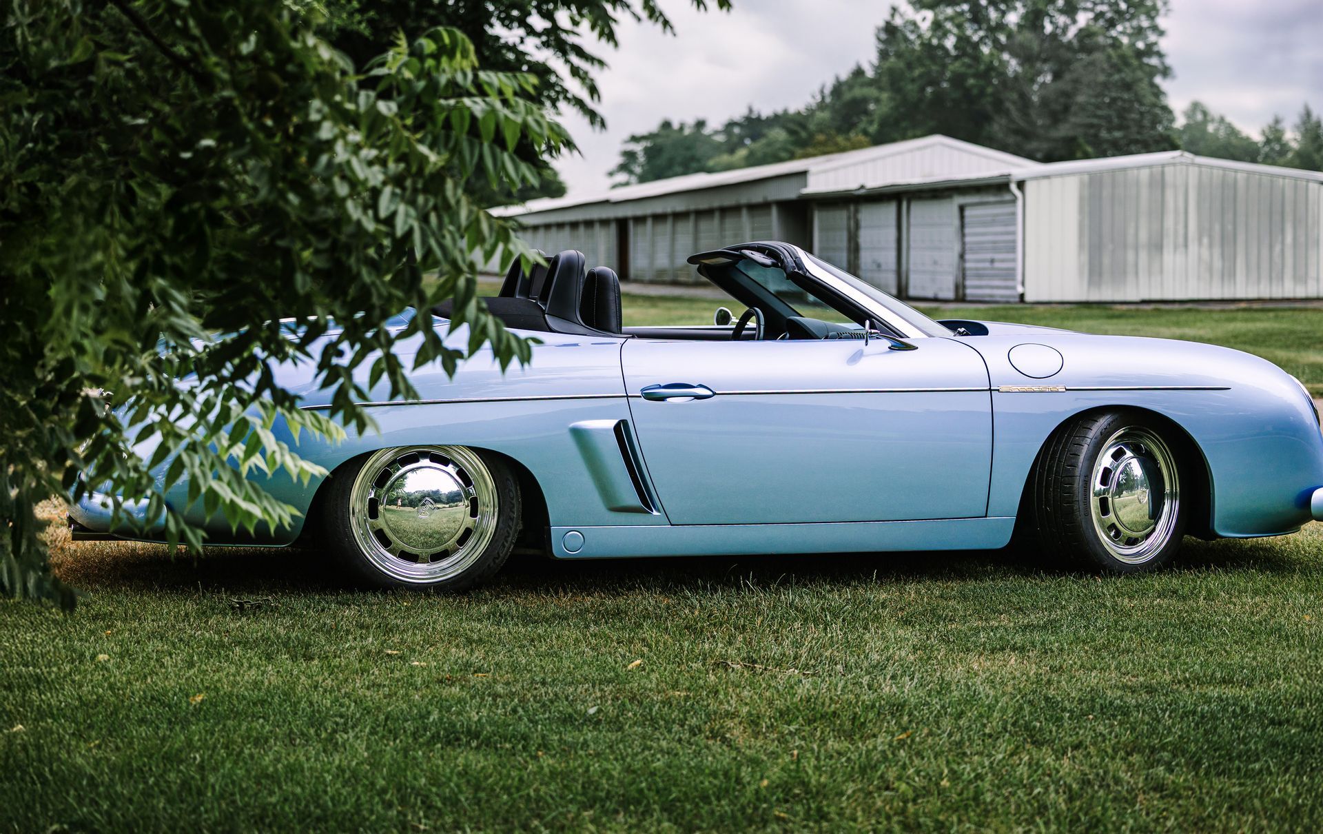 Light blue classic convertible parked on grass beside a white house and trees