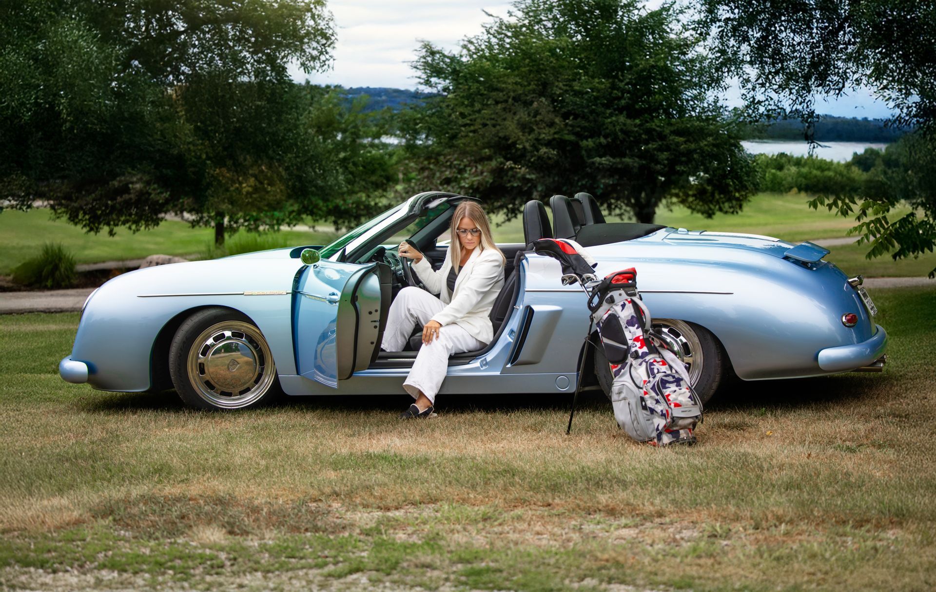 Woman sitting in a silver convertible on grass beside a floral suitcase, with trees and water behind.
