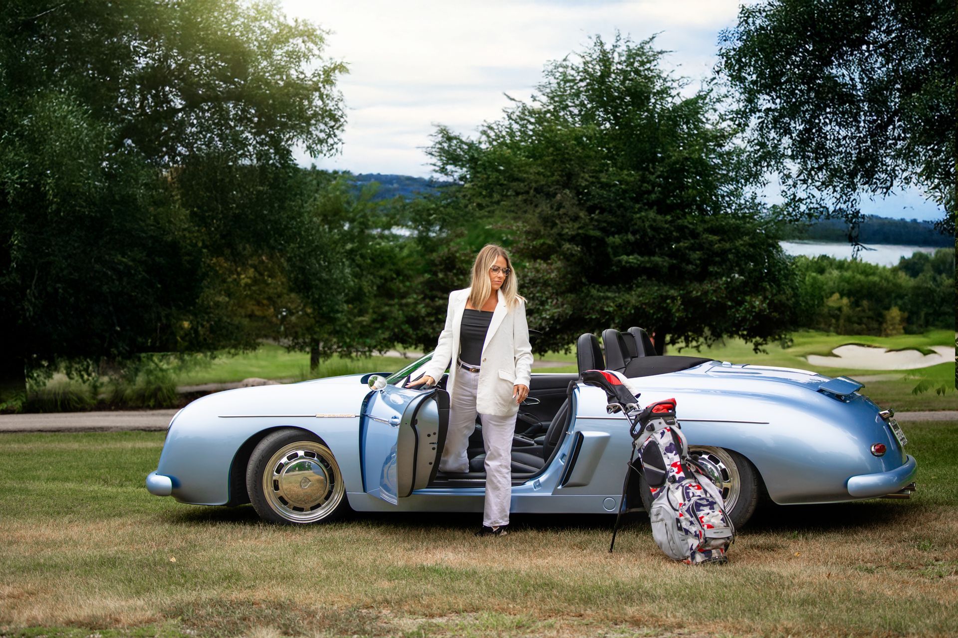 Woman standing by a light blue vintage car with an open door and floral bag on a grassy roadside.