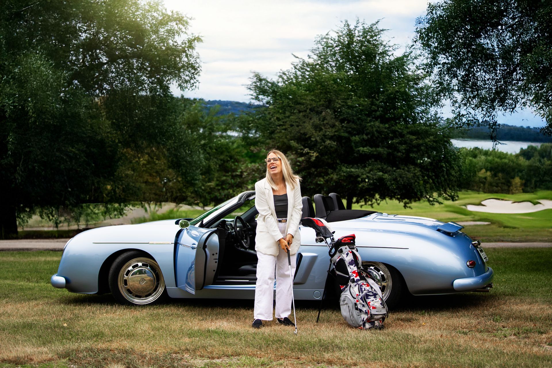 Woman in white beside a silver sports car with doors open, parked on grass near trees and water.