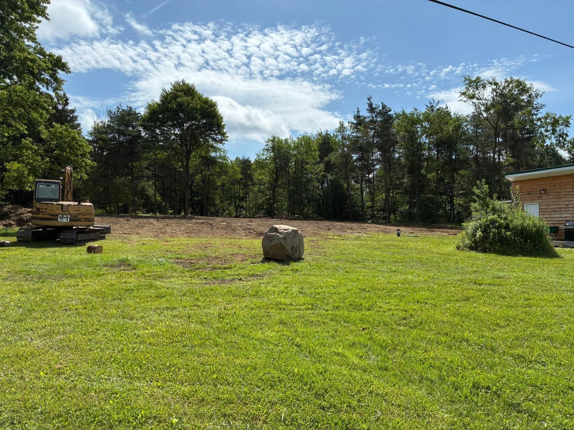 Grassy yard with a large rock, trees, a parked bulldozer, and a house under a blue sky