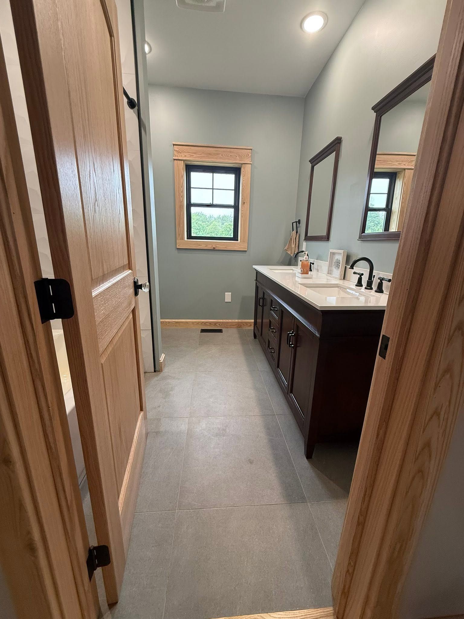 Narrow bathroom with double vanity, mirrors, gray tile floor, and a window at the far end.