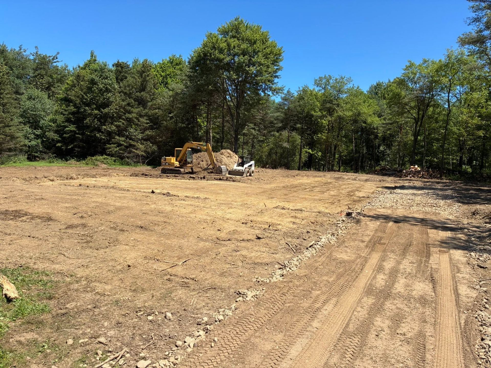 Cleared dirt lot with construction equipment beside a tree line under a bright blue sky
