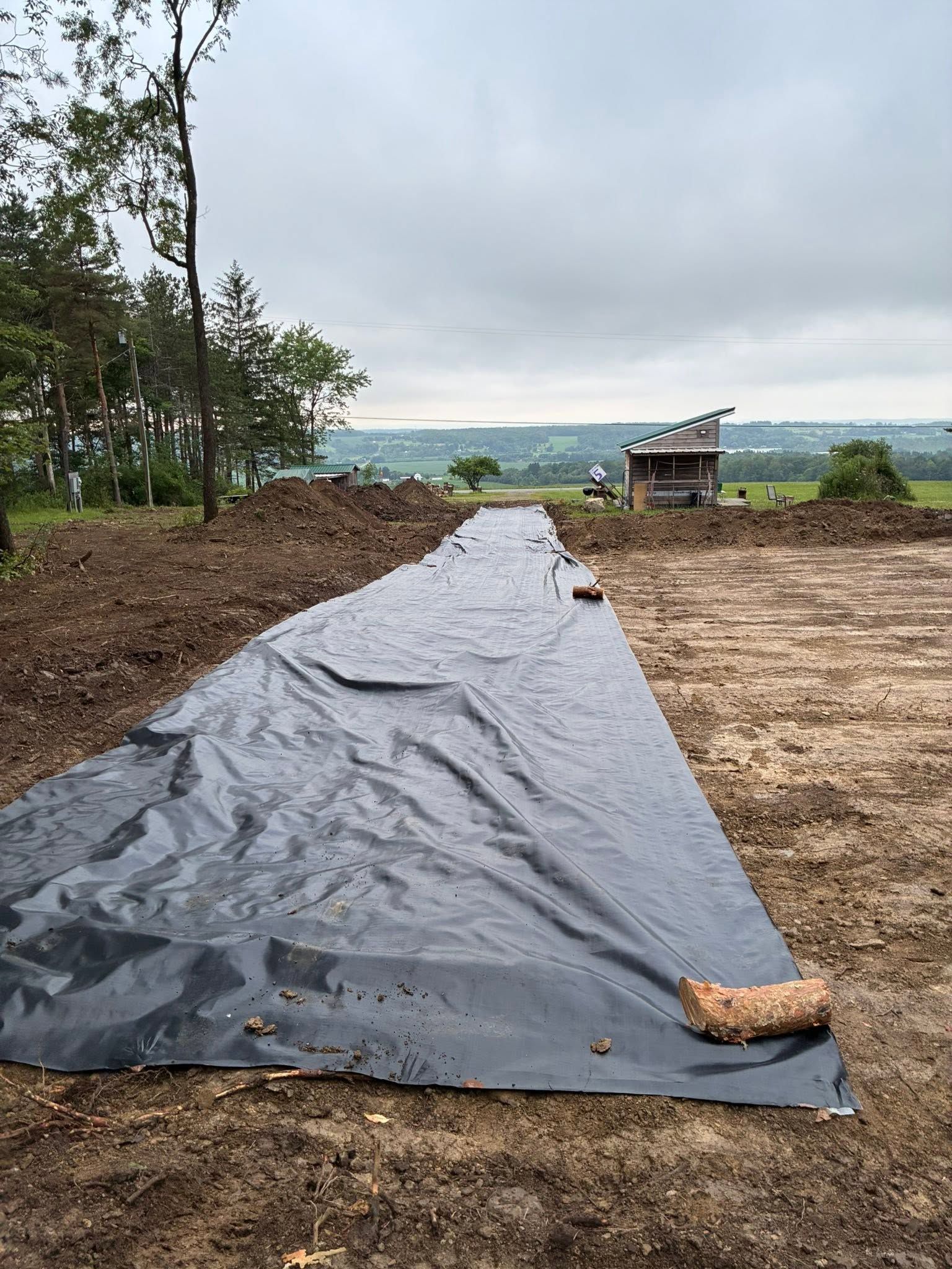 Black tarp-lined long plot on a dirt hillside under cloudy sky, with a small shed in the distance.