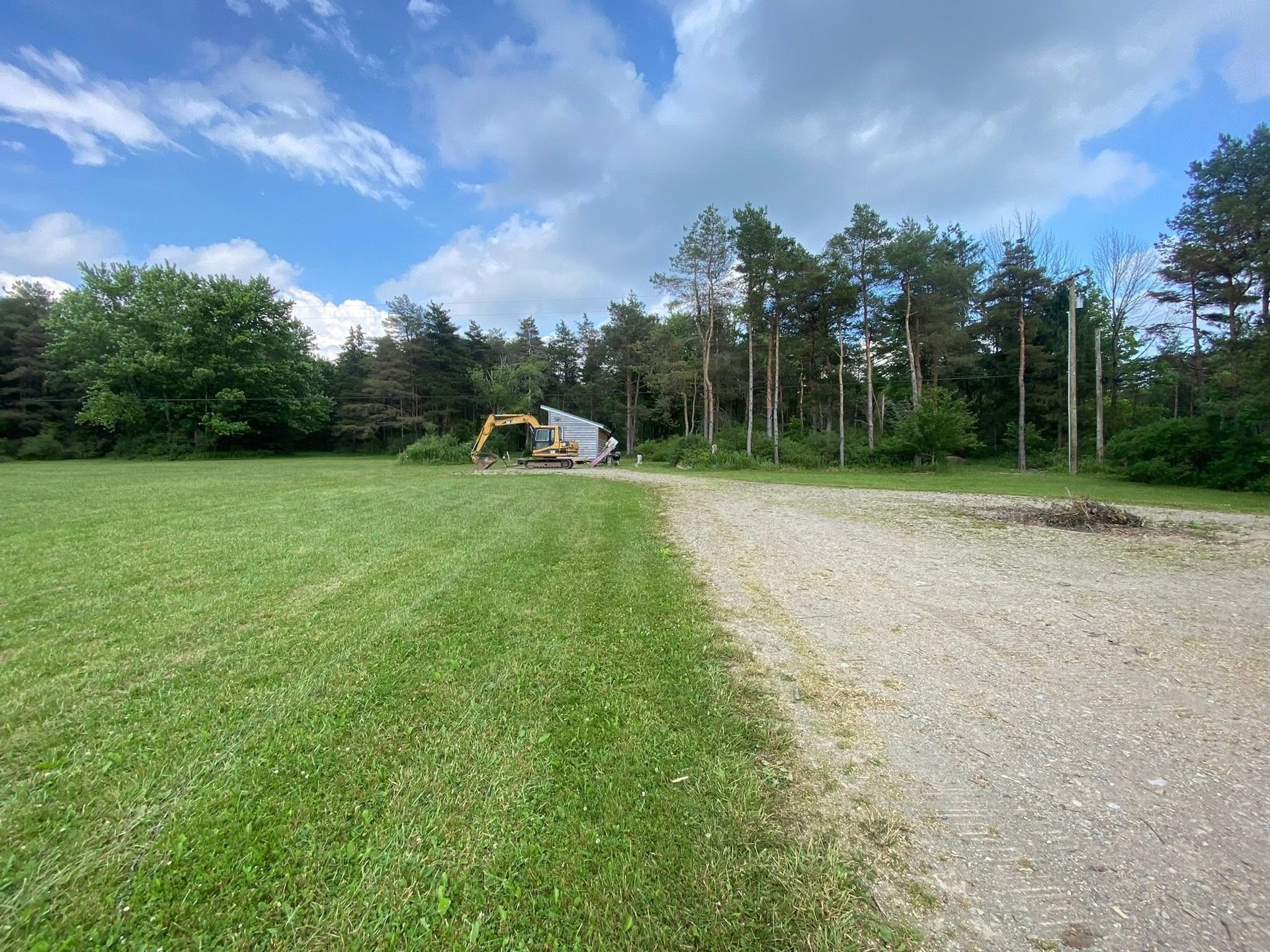 Gravel road beside a green field and trees under a blue sky, with a small excavator in the distance