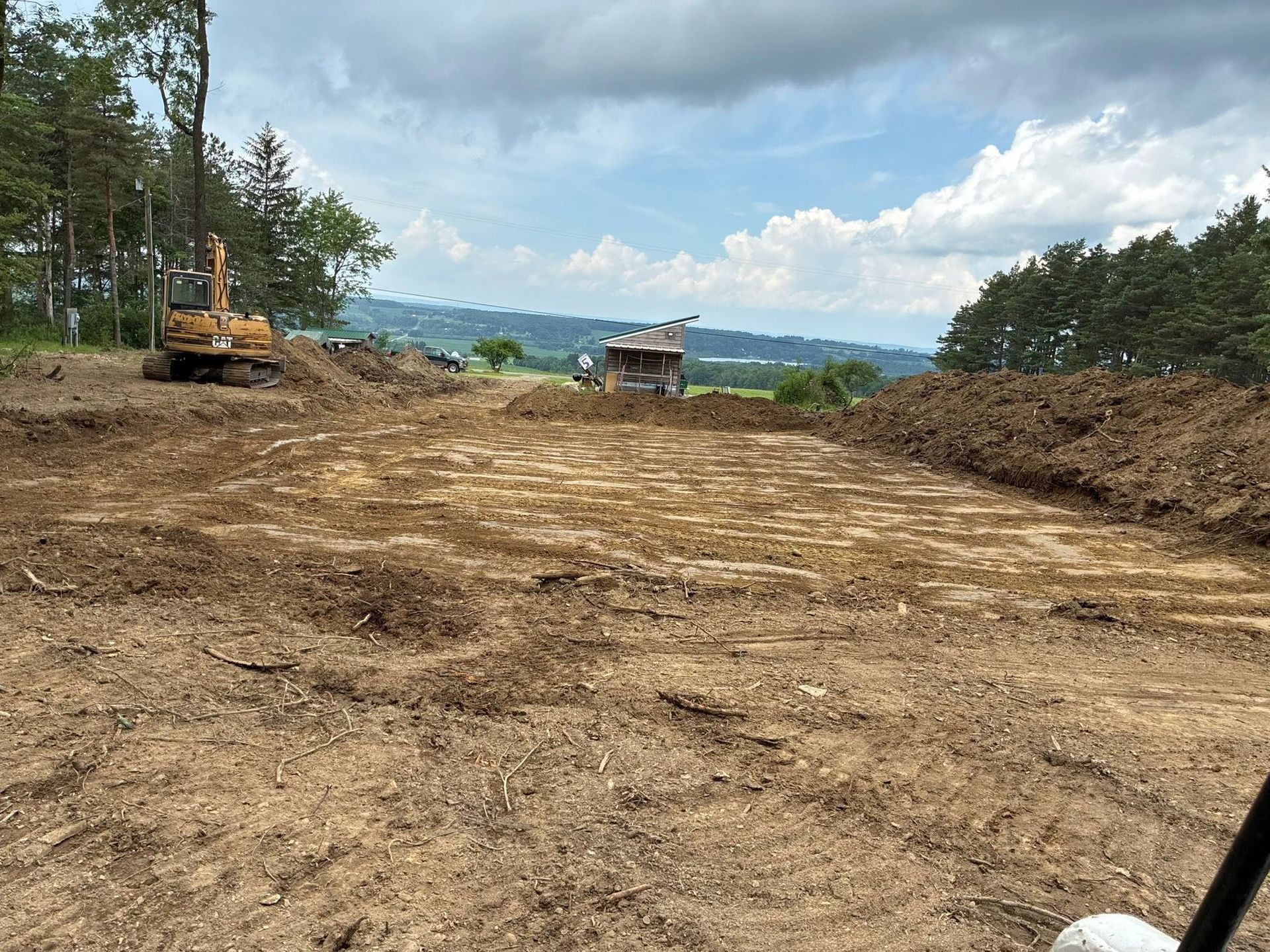 Cleared dirt road construction site with excavator, trees, and cloudy sky in the background