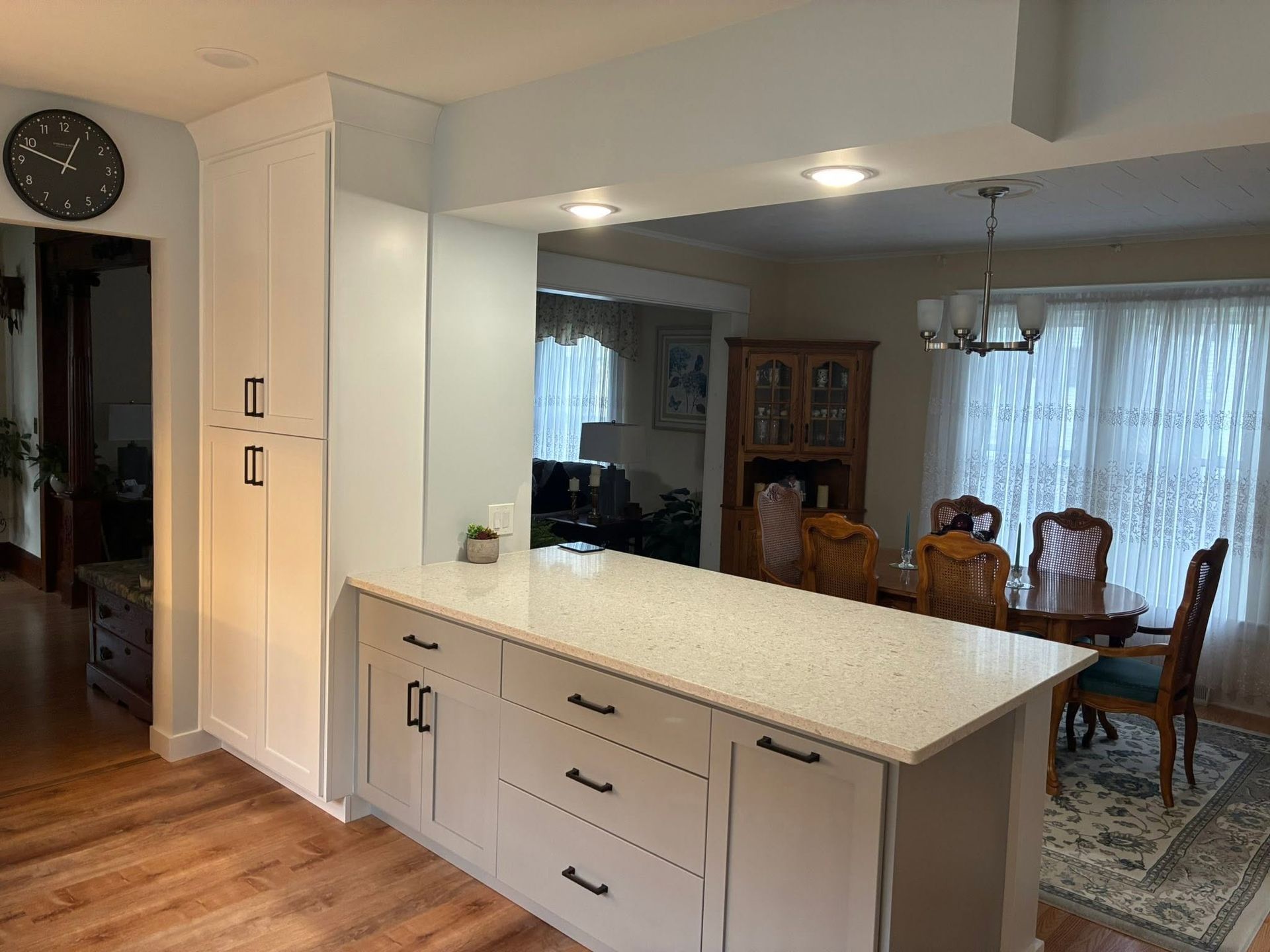 Bright kitchen with white island, wood floor, and dining room with table, chairs, and cabinet in the background
