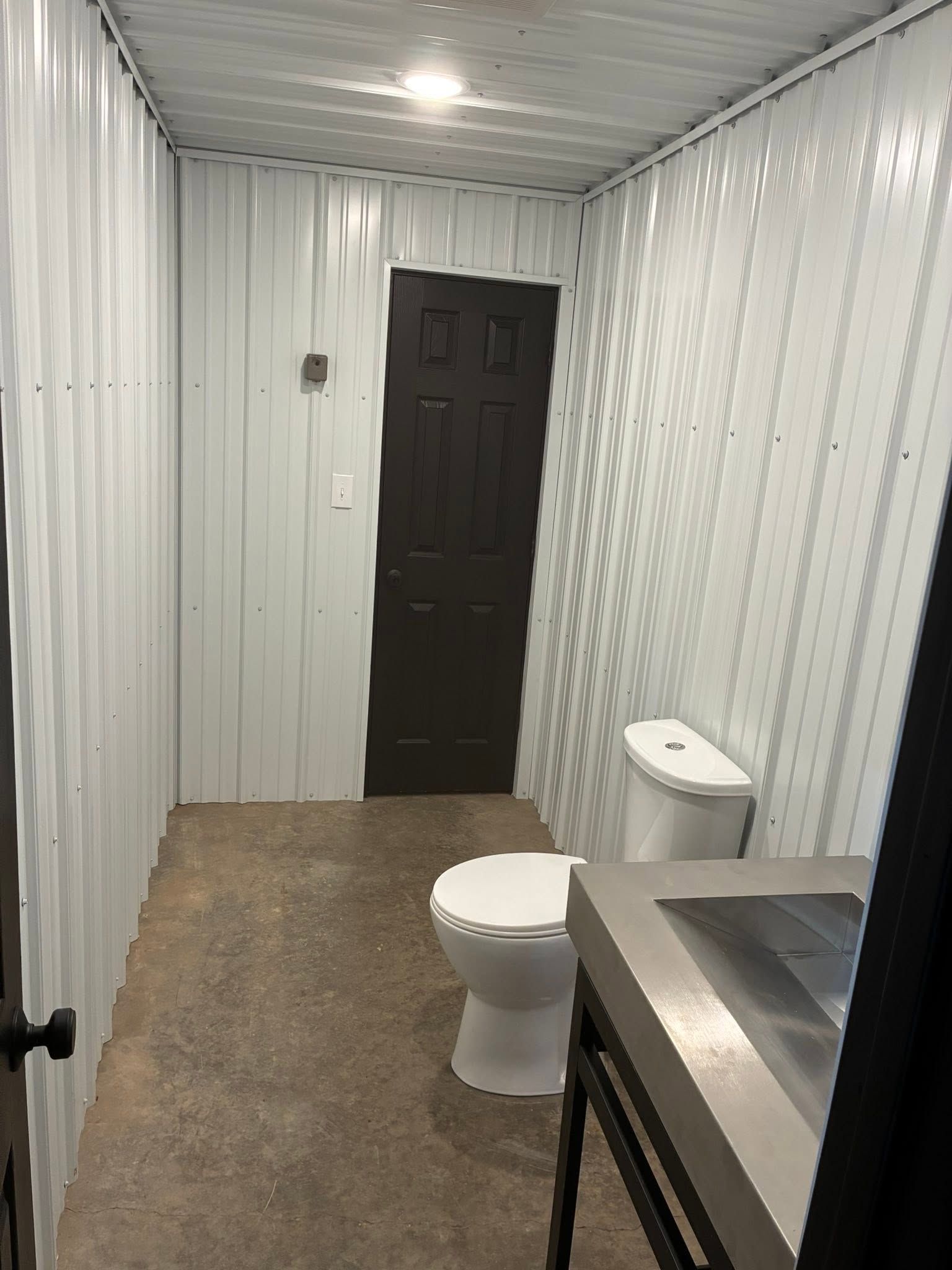 Small restroom with corrugated metal walls, toilet, sink, and brown door.
