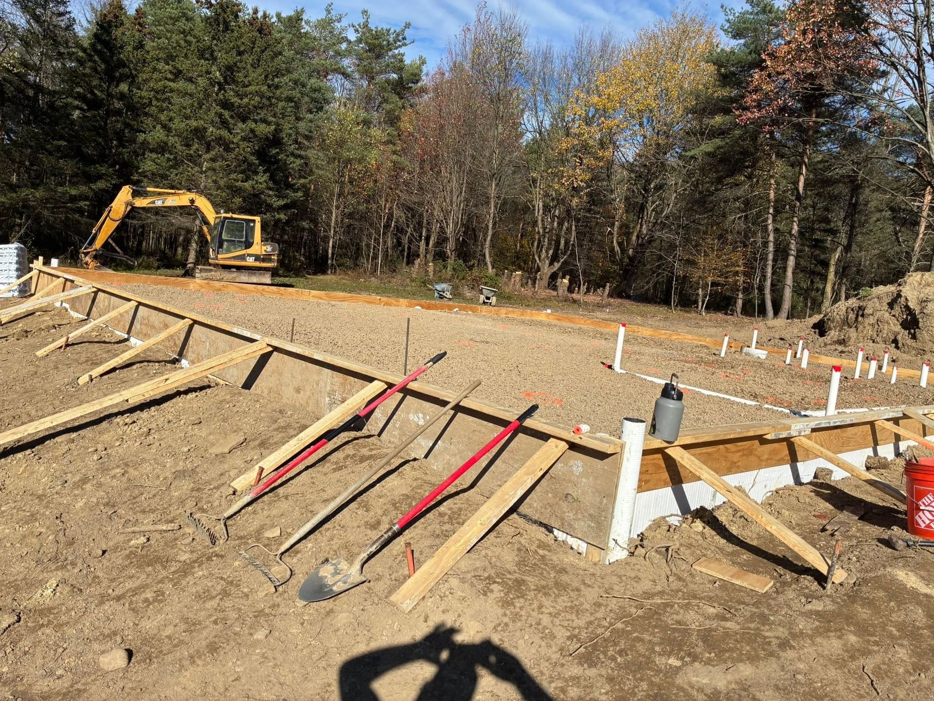 Construction site with excavator, dirt foundation, and white stakes outlining a building area in a wooded clearing