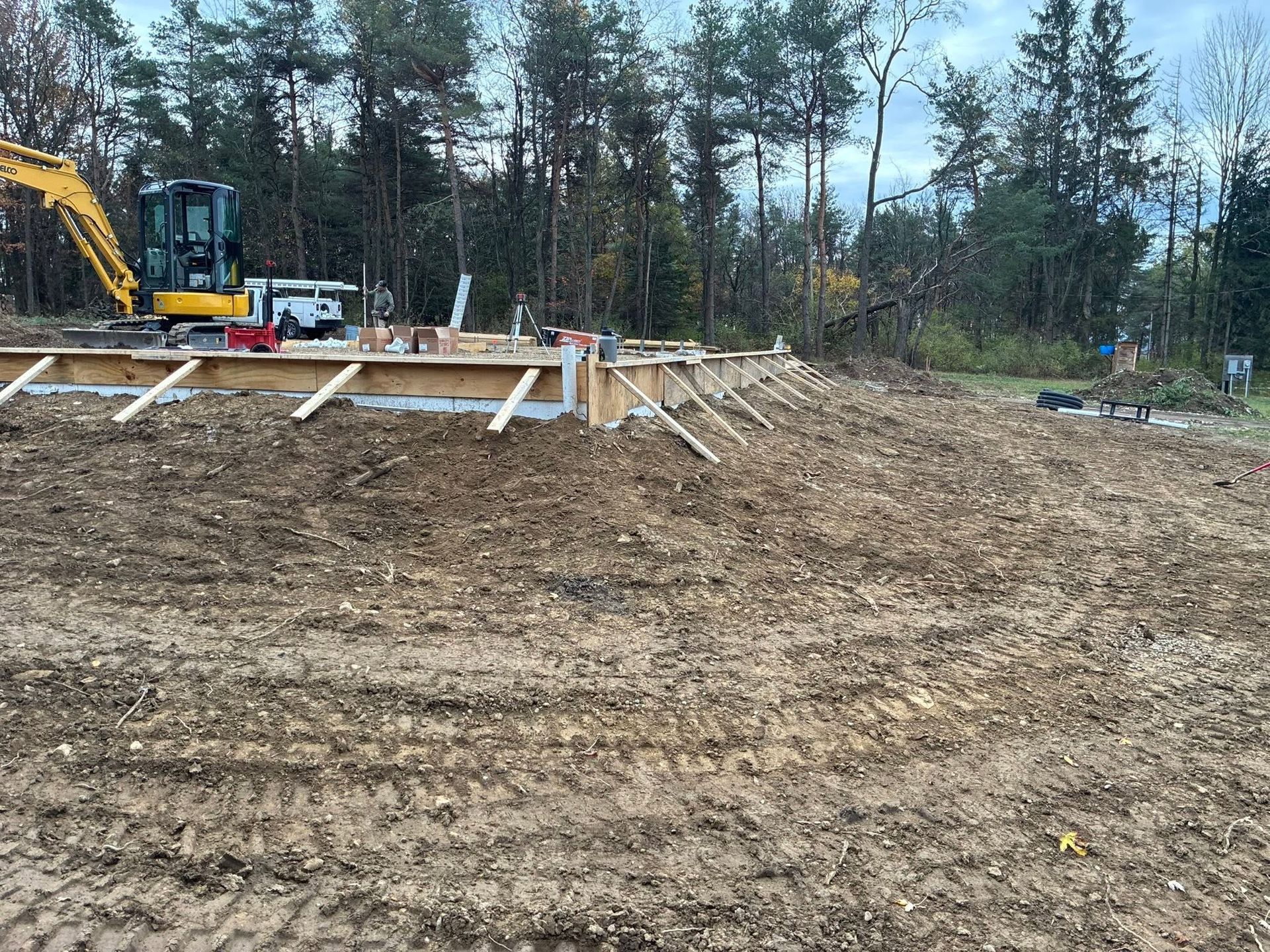 Construction site with excavator, wooden foundation forms, and a dirt clearing beside a wooded area