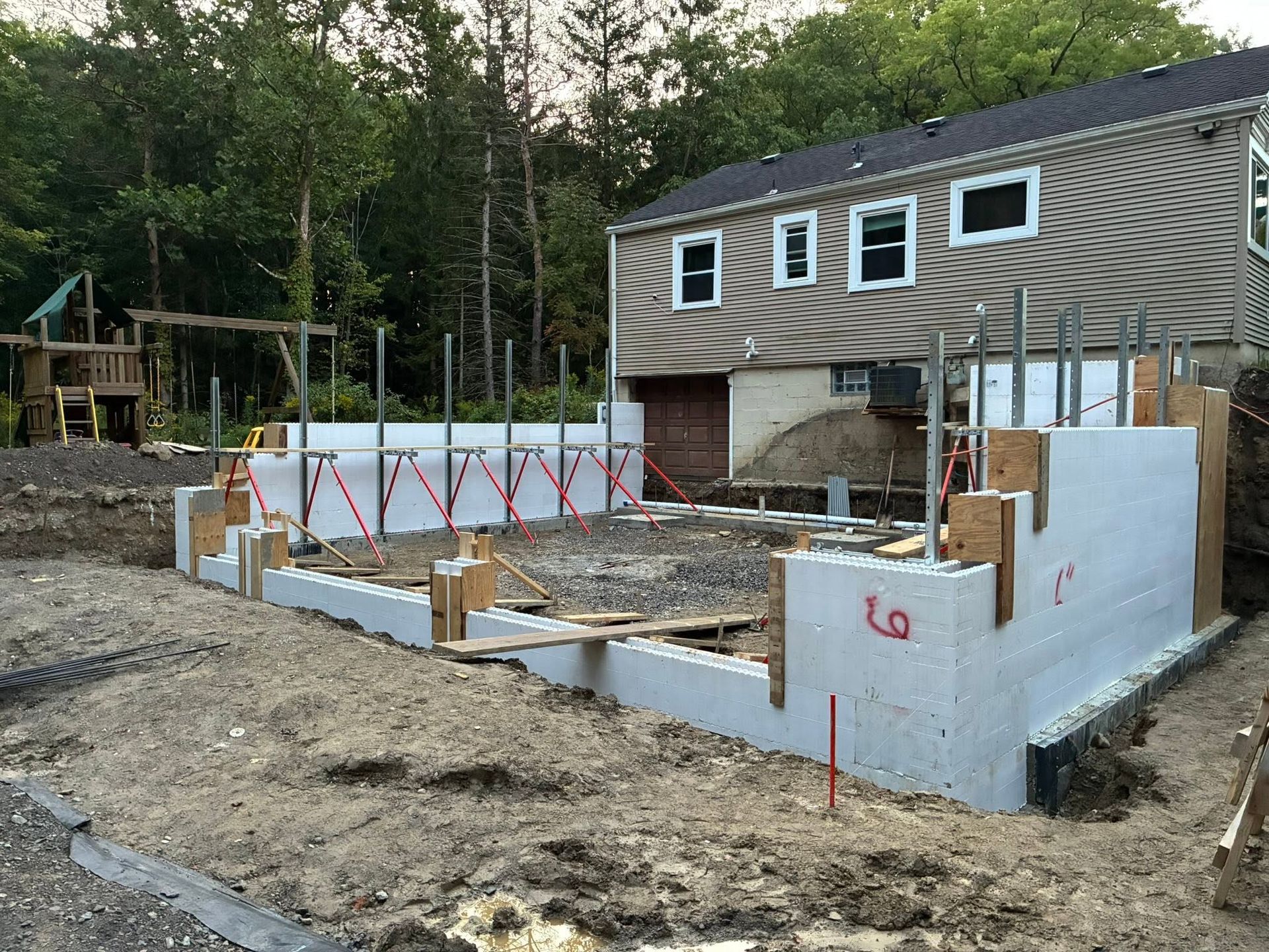 Concrete foundation construction with partial wood framing beside a house in a wooded area
